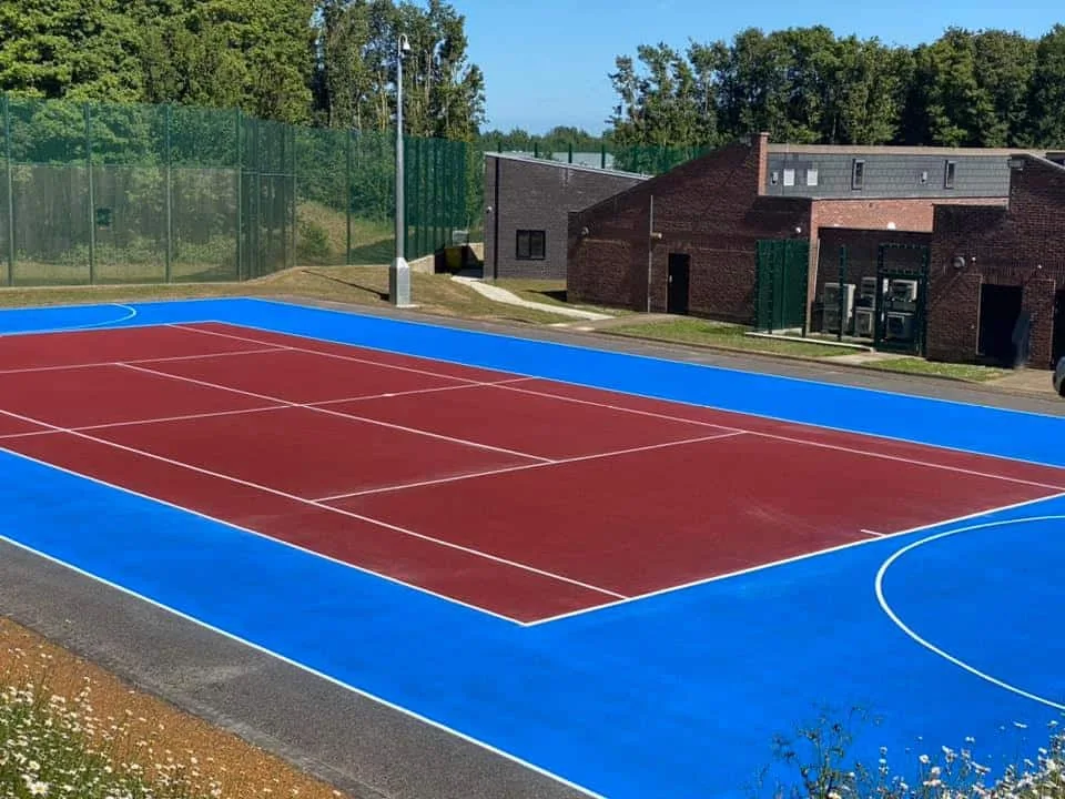A painted tennis court that has been painted on concrete with red, blue and white paint. In the distance there is a green fence and a brick building.