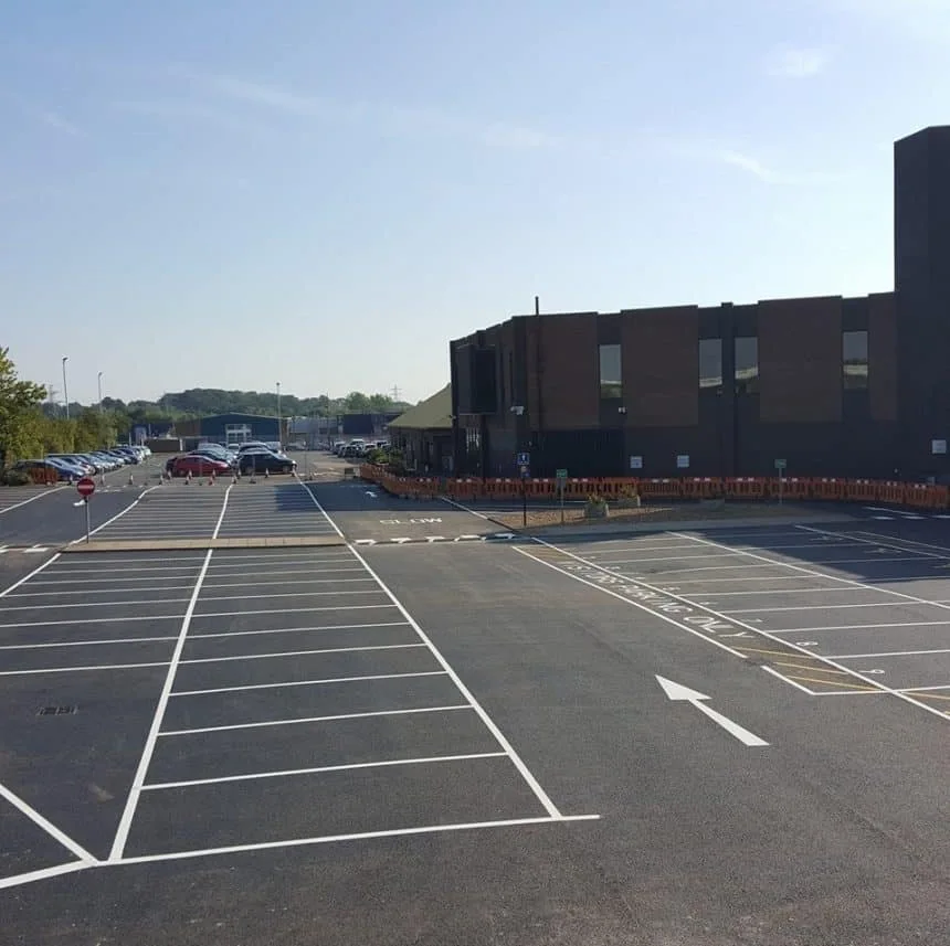 A car park with a one way system and no entry signs with white line painted parking bays. There is parked cars in the distance and orange barriers. Painted by line marking compaines.
