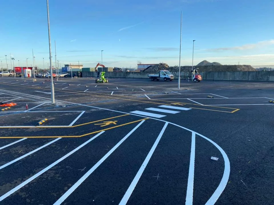 A car park that has been painted on tarmac with a yellow pedestrian walking zone and a zebra crossing. In the distance is a van with a cherry picker vehicle.