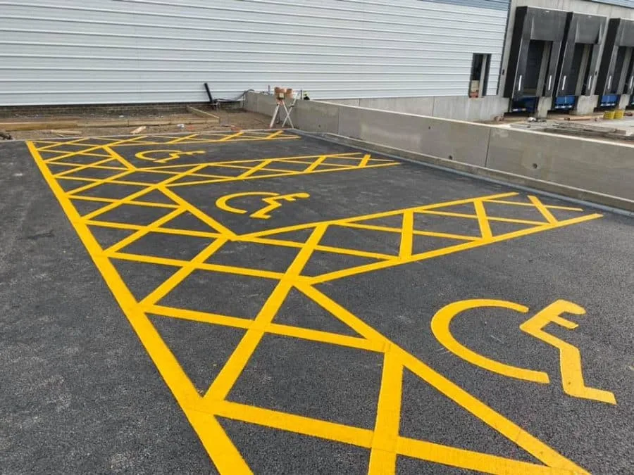 Yellow disabled car parking spaces that have been painted on tarmac in front of a wall near a building.