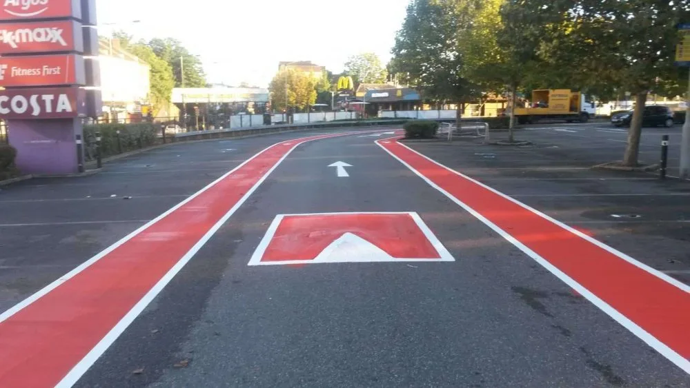 Red drive through road markings with a red speed bump in the middle and a car parking surrounding it. In the distance is fast food restaurants, trees and vehicles.