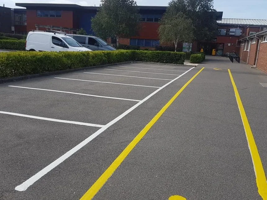 A small car park with a yellow line painted pedestrian walk way and two park vans in the distance as well as a building. painted by line marking companies